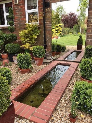 A brick bounded oblong pond with large mirror showing reflection of the garden