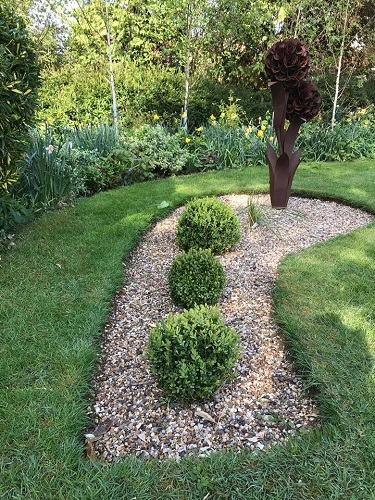 Oxbow shaped gravel bed with spherical planting and a tall metal flower like sculpture.