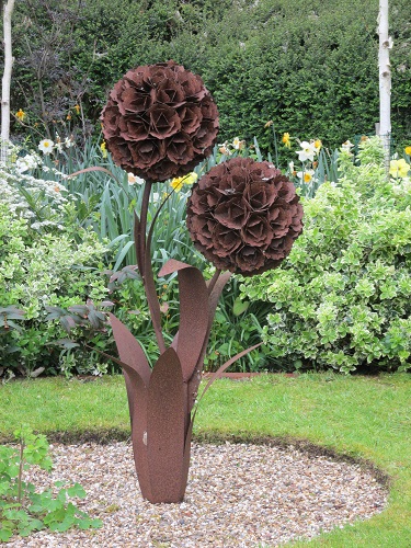 A rusty metal sculpture of a flower with 2 ball shaped blooms in a gravel border